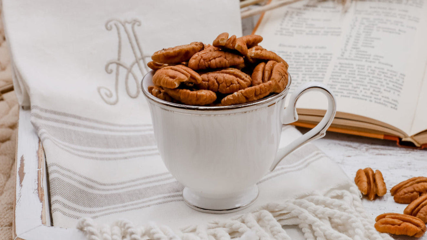 White teacup filled with pecans on a wooden surface with an open book and woven basket in the background.